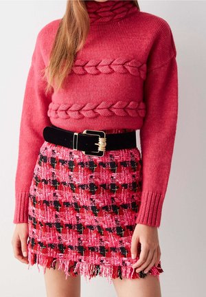 Pull en tricot rouge avec une texture tressée. Jupe courte en tissu tressé rose à franges avec un motif à carreaux et des accents noirs. Ceinture noire avec une boucle dorée.