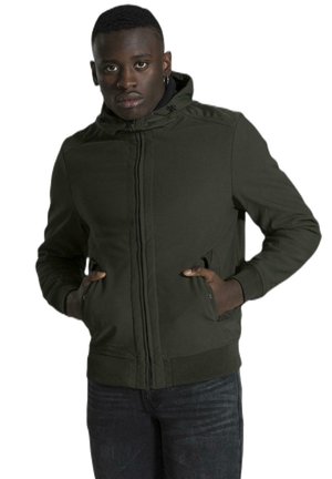 Un homme présente une veste à capuche vert olive foncé, les mains dans les poches, associée à un jean foncé sur fond blanc.