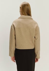 Giacca cropped marrone chiaro in un tessuto morbido e testurizzato, con un ampio colletto e spalle ribassate, indossata sopra un indumento nero.
