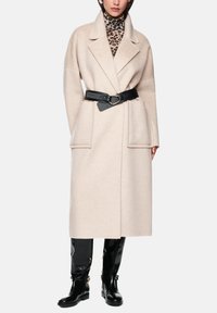 Long beige wool blend coat with wide lapels, a belted waist, and two large front pockets. Worn over a leopard print turtleneck and black boots.