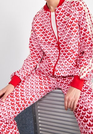 Persoon die een rood-wit gebloemd Adidas trainingspak met trefoil-logo's draagt, zittend op een metalen geribbeld oppervlak.