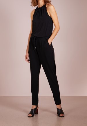 Schwarzer Jumpsuit mit gerafftem Ausschnitt, Kordelzug in der Taille und seitlichen Taschen, kombiniert mit schwarzen Sandaletten mit offenem Zeh und gemustertem Absatz.