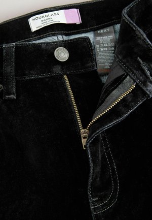 Schwarze Cordhosen mit hoher Taille, ausgestattet mit einem Metallknopf, Reißverschluss und Pflegeetikett, die eine glatte Textur und einen subtilen Glanz zeigen.