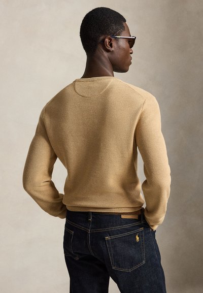 Uomo con maglione beige di maglia e jeans blu scuro in piedi con le mani nelle tasche posteriori, visto di profilo su uno sfondo semplice.