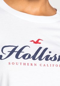 Vit bomull t-shirt med blå kursiv "Hollister" text och röd "SOUTHERN CALIFORNIA" under, med en liten röd måslogotyp.