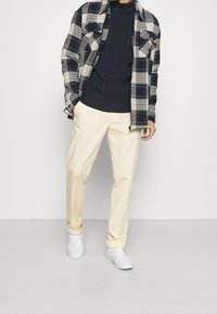 Maglione lavorato a maglia a coste blu marina sotto una camicia a quadri bianca e nera, abbinato a pantaloni giallo chiaro e sneaker bianche. Trame morbide ovunque.