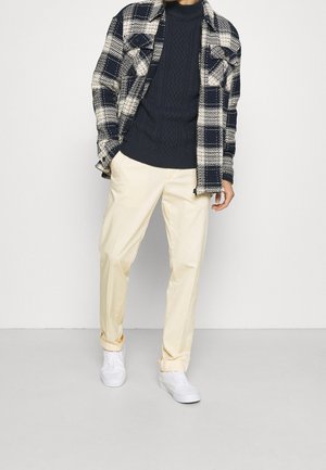 Suéter de punto trenzado azul marino debajo de una camisa de cuadros en blanco y negro, combinado con pantalones amarillo claro y zapatillas blancas. Texturas suaves en todo el conjunto.