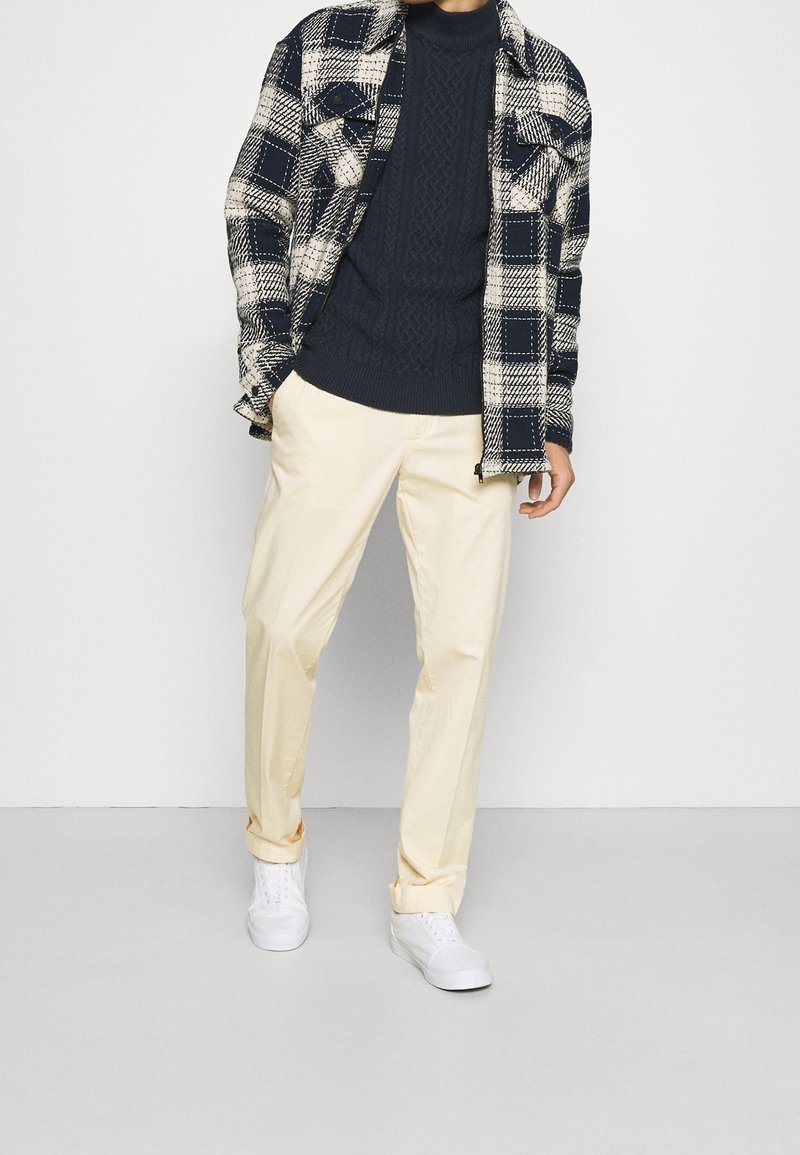 Maglione lavorato a maglia a coste blu marina sotto una camicia a quadri bianca e nera, abbinato a pantaloni giallo chiaro e sneaker bianche. Trame morbide ovunque.