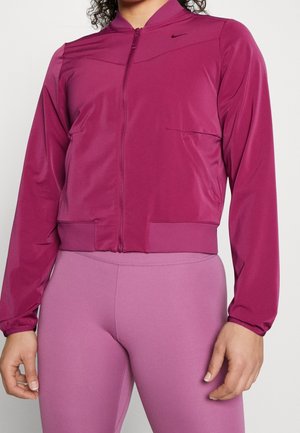 Kvinna iklädd en åtsittande magenta zip-up jacka och ljuslila högmidjade leggings, står med armarna avslappnade vid sidorna.