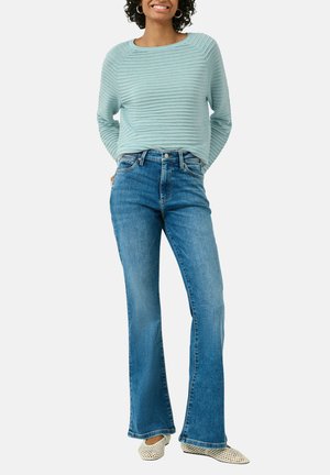 Femme souriante portant un pull côtelé bleu clair, un jean bleu évasé taille haute et des chaussures plates blanches tissées, debout avec les mains dans le dos.