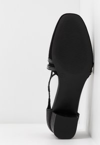 Chaussure pour femme noire à talon carré vue de la semelle, inclinée contre un mur d'angle blanc et un sol.