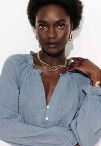 Blouse bleu clair à manches longues en tissu texturé, dotée d'un col et de poignets volantés, avec un devant à boutons et une coupe décontractée.