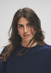 Mujer con cabello largo y oscuro, vistiendo un suéter de canalé azul marino, mirando directamente a la cámara con una expresión neutral sobre un fondo claro y liso.