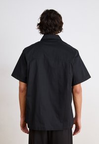 Camicetta nera a maniche corte con colletto classico, realizzata in tessuto liscio, caratterizzata da un taglio dritto e dettaglio della yoke posteriore.