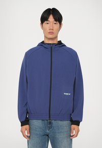 Violet zip-up striukė su kapišonu, su juodais akcentais ant rankovių ir apatinio krašto, bei mažas akvamarino spalvos logotipas priekyje. Lygus, lengvas medžiaga.