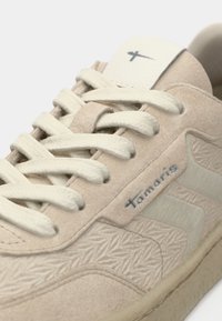 Beige Sneaker mit strukturiertem Wildleder und glatten Akzenten. Verfügt über cremefarbene Schnürsenkel, ein prominentes Logo und ein gemustertes Design an den Seiten.