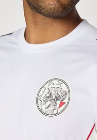 Maillot de sport blanc avec un logo Ajax rond présentant le profil d'une figure et des accents rouges sur les épaules. Texture de tissu lisse.