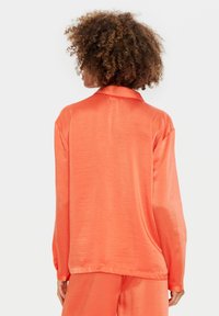 Blusa a maniche lunghe di un brillante arancione, realizzata in tessuto morbido. Presenta un colletto classico e una vestibilità rilassata, con dettagli di cucitura visibili.