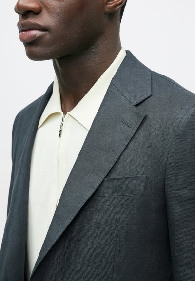 Κοντινή λήψη ενός άνδρα που φοράει σκούρο γκρι σακάκι πάνω από μια κρεμ ζακέτα με φερμουάρ, εμφανίζοντας μέρος του προσώπου και του άνω κορμού του.