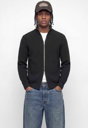 Jeune homme aux cheveux tressés portant un pull zippé foncé, une chemise blanche, un jean bleu et une casquette de baseball sombre, debout les mains dans les poches.