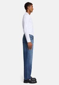 Jeans in denim blu medio, taglio dritto, con due tasche posteriori e dettagli di cucitura, indossati con una camicia bianca a maniche lunghe e scarpe nere.