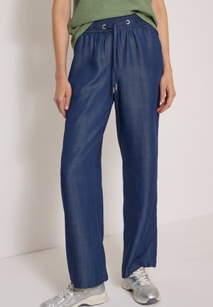 Kvinde iført løstsiddende blå denim bukser med elastisk talje og snøre, kombineret med sølvfarvede sportssko og en grøn top.