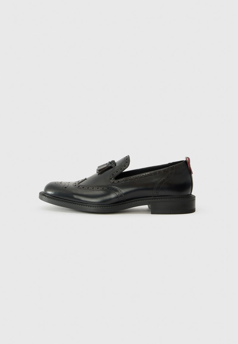Black leather loafers with brogue detailing, featuring a flat sole and decorative hardware at the front. Smooth texture and minimalistic design.