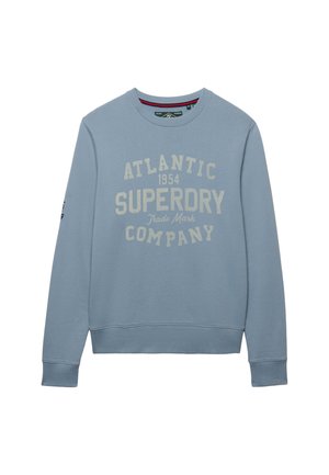 Lyseblå crewneck-sweatshirt med lange ærmer og hvid tekst "Atlantic 1954 Superdry Trade Mark Company" på forsiden.