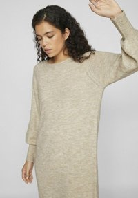 Beige stickad klänning med lång, lätt pösig ärm. Mjuk textur och avslappnad passform, med rund halsringning och ribbade muddar.