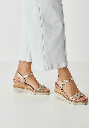 Keilsandalen mit einem metallisch goldenen Riemen und geflochtenem Vorderteil, einem korkähnlichen Absatz und einer robusten Gummisohle, entworfen für Komfort und Unterstützung.