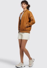 Giacca bomber marrone con polsini e orlo a costine, presenta una zip frontale e lettering ricamato. Indossata con pantaloni corti bianchi e sneakers beige.