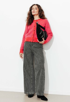 Femme portant un cardigan rouge boutonné, un jean gris à jambes larges, des bottes noires, tenant un sac à main noir avec des œillets argentés sur un fond blanc.