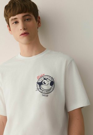 Young man wearing a white T-shirt with a winking black cartoon cat head and the words "Felix the Cat" on the chest.
