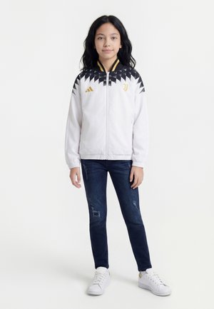 Pige iført hvid og sort sportjakke med guld logoer, mørke revnede jeans og hvide sneakers, stående mod en hvid baggrund.