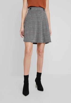 Kobieta ubrana w brązowy top, szarą kratkowaną spódnicę do połowy uda oraz czarne, wysokie botki na obcasie, stojąca na tle jednolitego szarego tła.