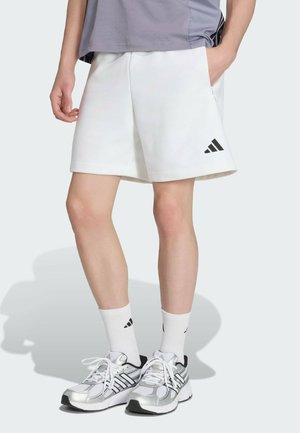 Personne portant un short blanc Adidas, des chaussettes blanches Adidas et des chaussures de course argentées Adidas, debout avec une main dans la poche.