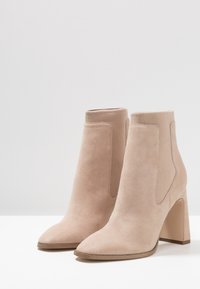 Botines beige de ante con punta afilada y paneles laterales elásticos. Cuentan con un tacón grueso de 3.5 pulgadas y una textura suave.