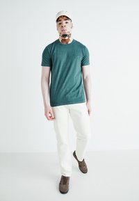 Teal kortermet t-skjorte med teksturert overflate og preget logo, kombinert med kremfargede slim-fit bukser og brune snøringer.