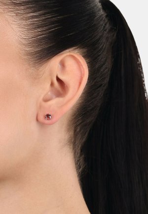 Primer plano de la oreja de una mujer con el cabello oscuro recogido, que lleva un pequeño pendiente redondo con una piedra preciosa roja.