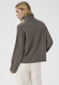 Gray knit sweater with a high collar, relaxed fit, and ribbed hem. The fabric appears soft and textured, contrasting with light-colored pants.