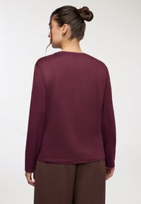 Top bordeaux a maniche lunghe con scollatura a girocollo, texture liscia, vestibilità rilassata e dettagli di cucitura minimi, abbinato a pantaloni larghi marroni.