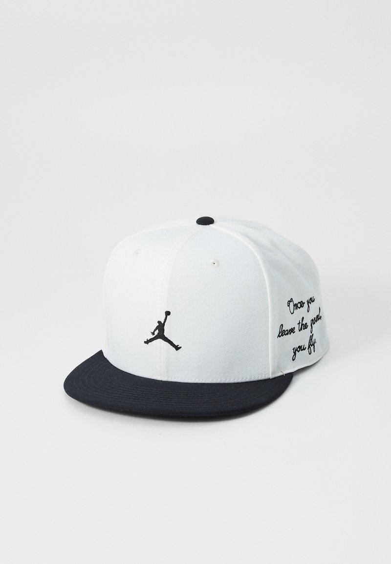 Balta cepure ar melnu vizieri un izšūtu Jumpman logo; uz sāniem ir teksti, izgatavota no auduma ar strukturētu piegriezumu.