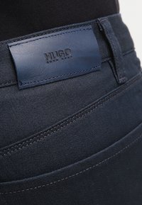 Mörka denimjeans med en slät yta, kontrasterande sömmar och en blå läderlapp präglad med "HUGO" på midjan.