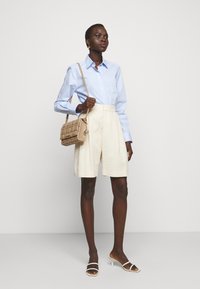 Camicia button-up azzurro chiaro, pantaloni corti crema su misura, borsa trapuntata beige e sandali bianchi con fasce. Texture lisce e linee pulite.