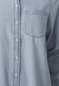 Light blue denim shirt with white buttons and a single chest pocket on the left side, shown in close-up view.