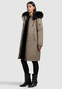 Pikk khaki parka eemaldatava musta karva kapuutsiga, lukuga taskutega ja tikanditakke tekstuuriga. Kombineeritud musta kleidi ja retuusidega.