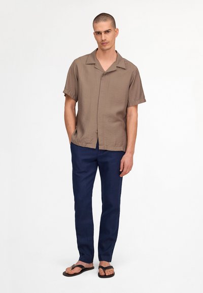 Homme portant une chemise marron à manches courtes boutonnée, un pantalon bleu marine et des tongs noires, debout devant un fond blanc uni.