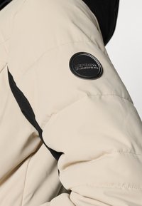 Manga de uma jaqueta puffer bege com detalhes em preto e um emblema redondo da marca com texto em relevo. O tecido parece suave e texturizado.