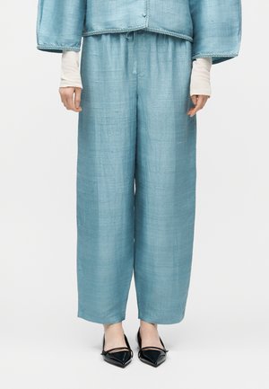 Laisvai krentančios šviesiai mėlynos tekstūruotos kelnės, derančios su tokiu pačiu šviesiai mėlynu švarku, ir juodi bateliai su smailiomis nosytėmis ant žmogaus, kurio rankos ramiai nuleistos šonuose.