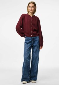 Burgunderfärgad stickad kofta med rund halsringning och vita knappar, i kombination med ljusblå utsvängda jeans och beige skor, bärs av en modell.
